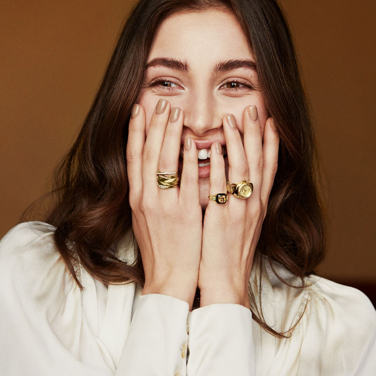An image of a woman wearing gold-tone rings and covering her smile. An image of the gold-tone Watch Ring with a green malachite dial.