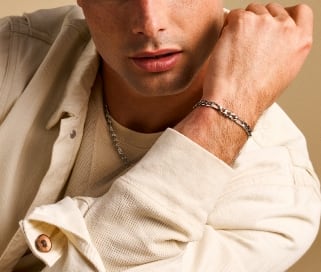 Un hombre con una camisa en tono marfil que lleva un collar y una pulsera en tono plateado.